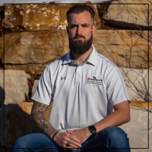 An alert white man with short brown hair and a beard sits with his hands in his lap