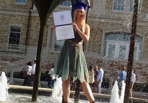 A young woman w long, blond hair stands near fountains in a blue graduation cap