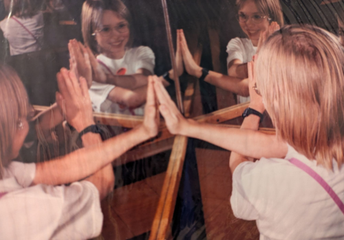 A blond girl places each hand on a separate mirror & views her multiple reflections