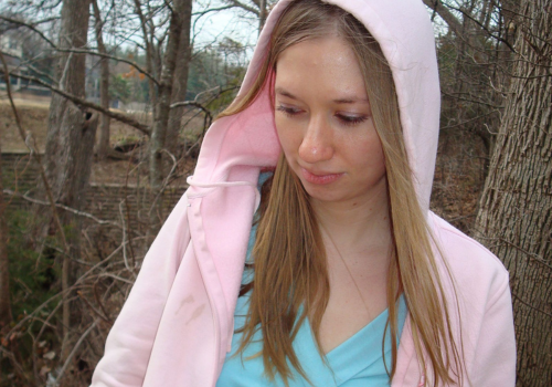 A young woman with long, blonde hair wears a pink hoodie and looks down, away