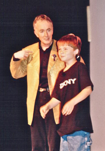 An older white man in a gold blazer & a white boy in a black t-shirt enter a stage