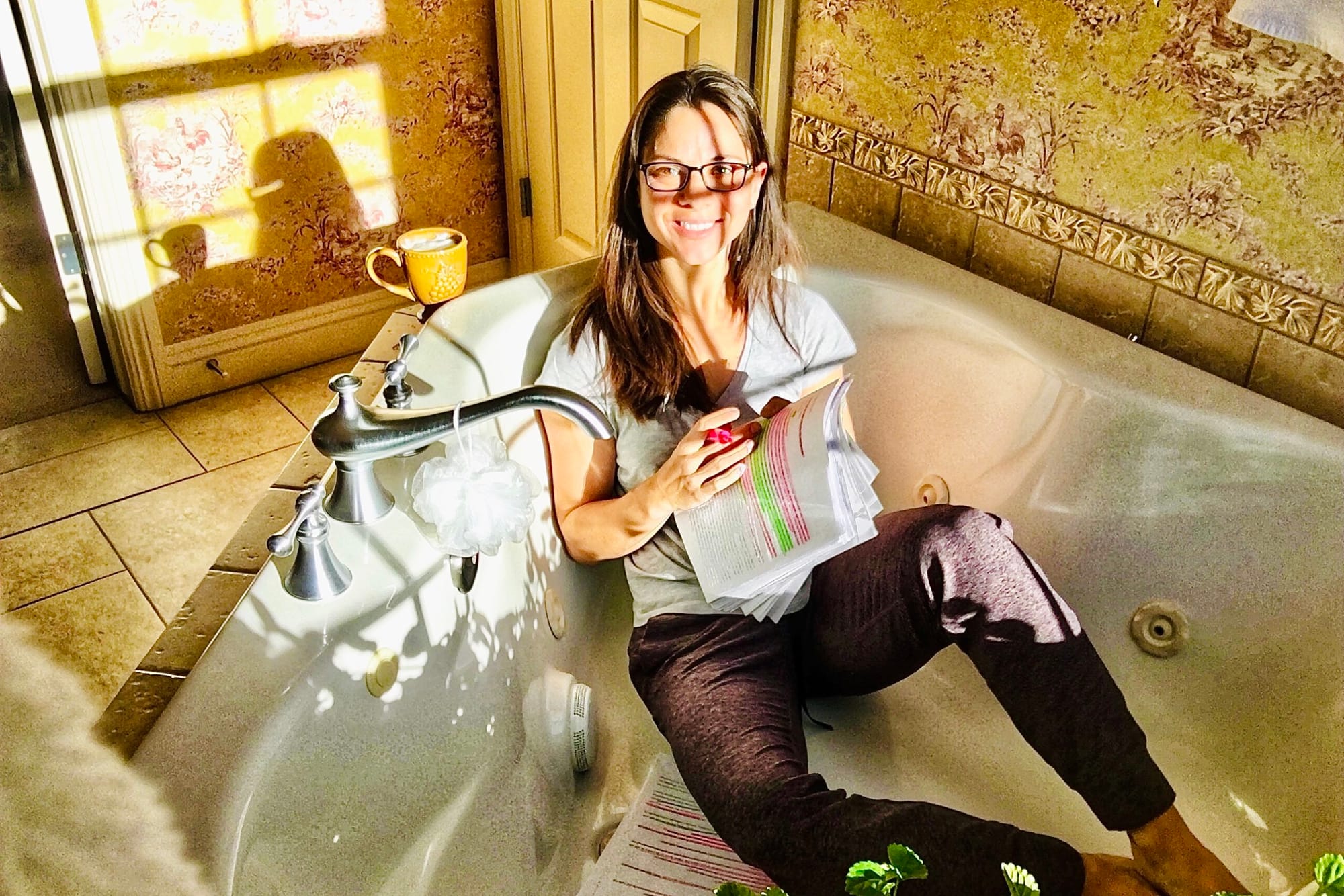 Woman w/shoulder-length brown hair and wearing glasses sits in an empty bathtub