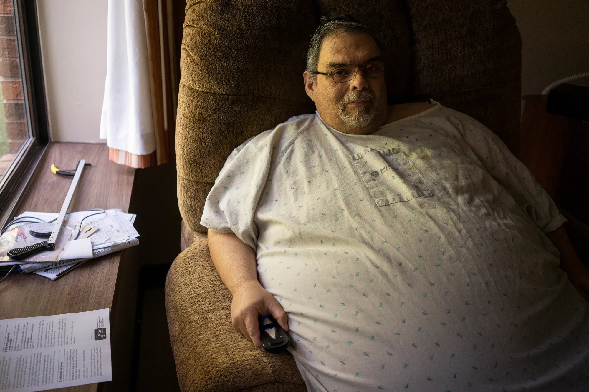 An obese man w/short, iron gray hair & wearing glasses sits in a chair look at a TV