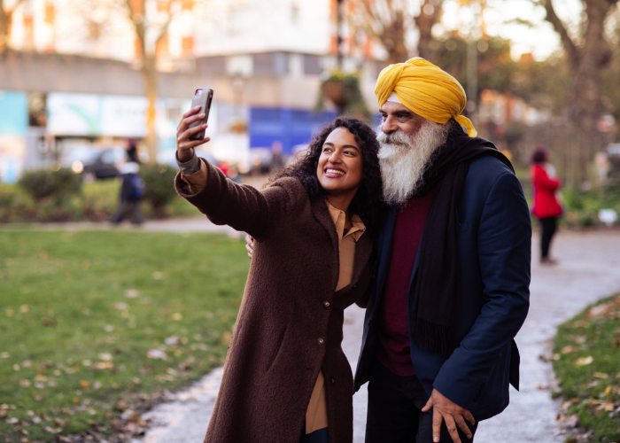 A Sikh man witha flowing, white beard & wearing a gold turban takes a selfie w/a young woman with curly, brown hair in a par