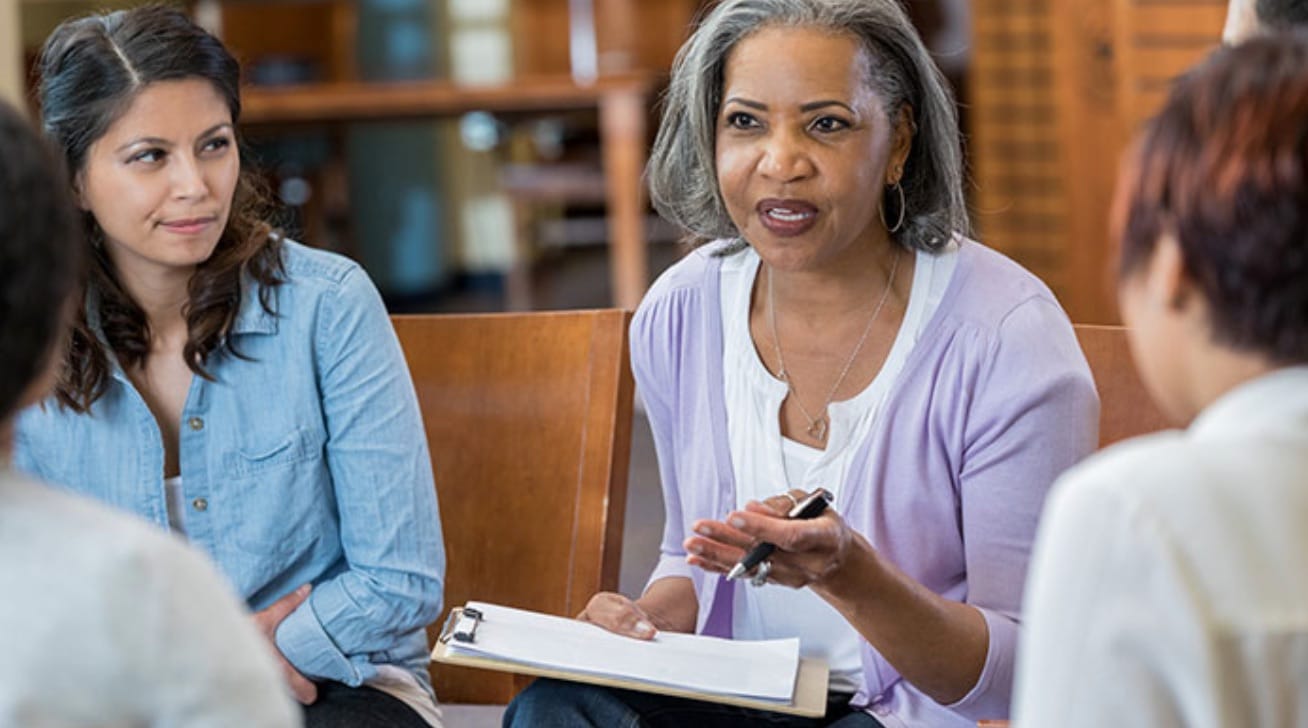 A middle-aged Black woman w/a clipboard speaks to a women's group. A Latina and others listen