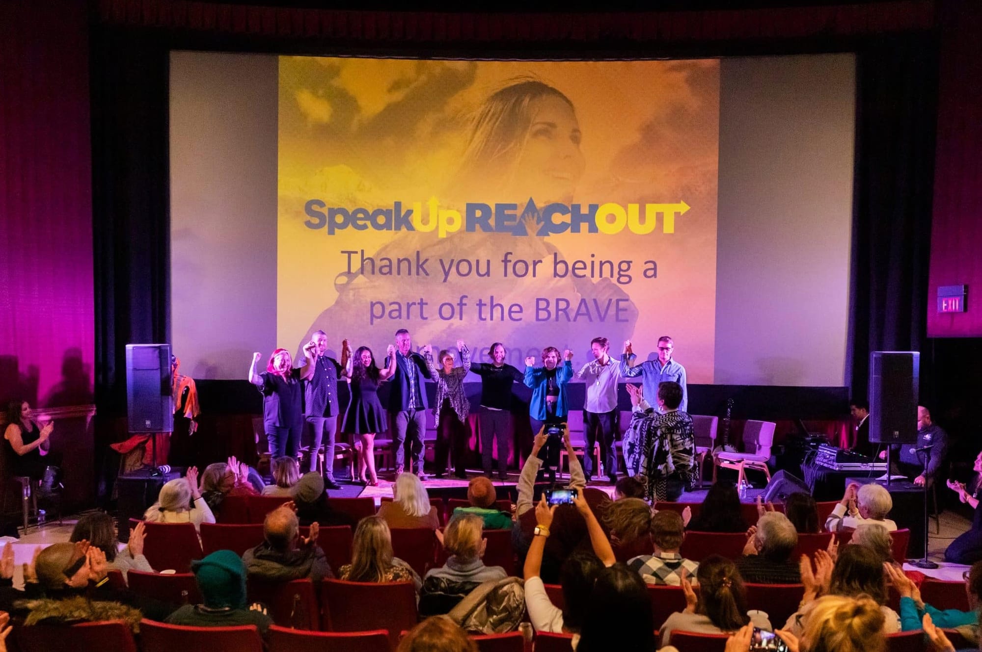 Photo of a theater. 9 people on stage holding up each other's hands. Audience clapping. SpeakUp ReachOut logo above stage