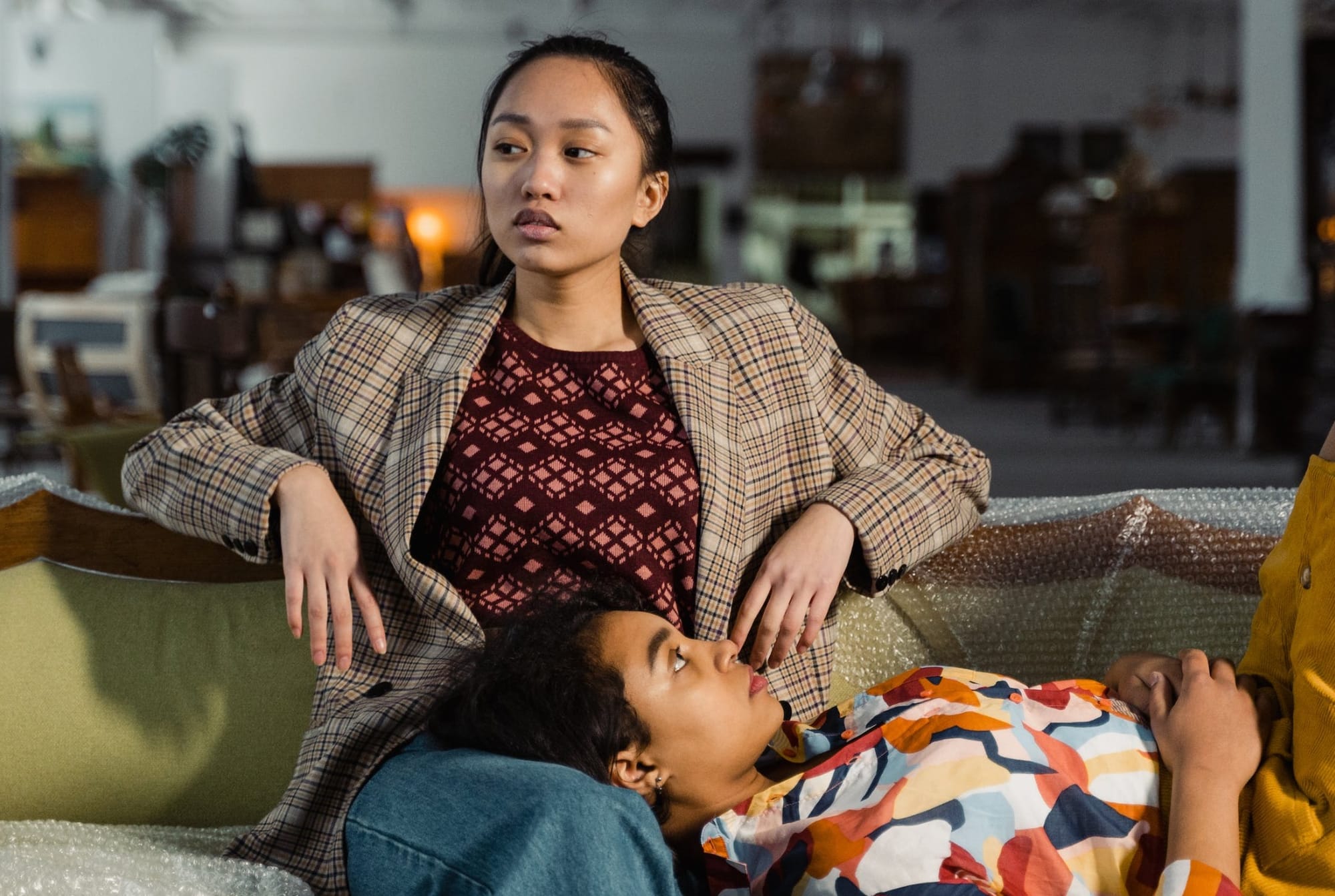 One woman sits on a couch in a large, cluttered room. Another woman lies with her head in the 1st woman's lap