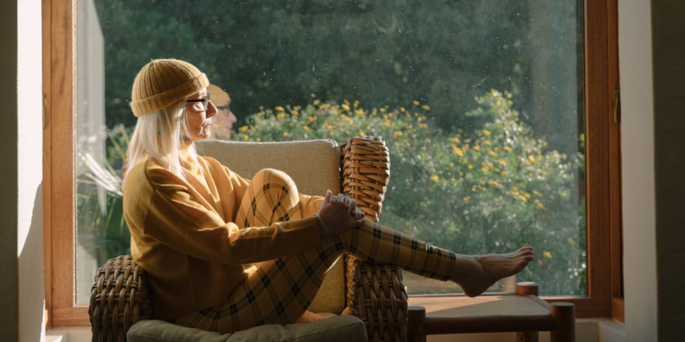 Older woman w/long, white hair sits sideways in a chair w/1 knee up & other leg extended over arm; she's looking out a window