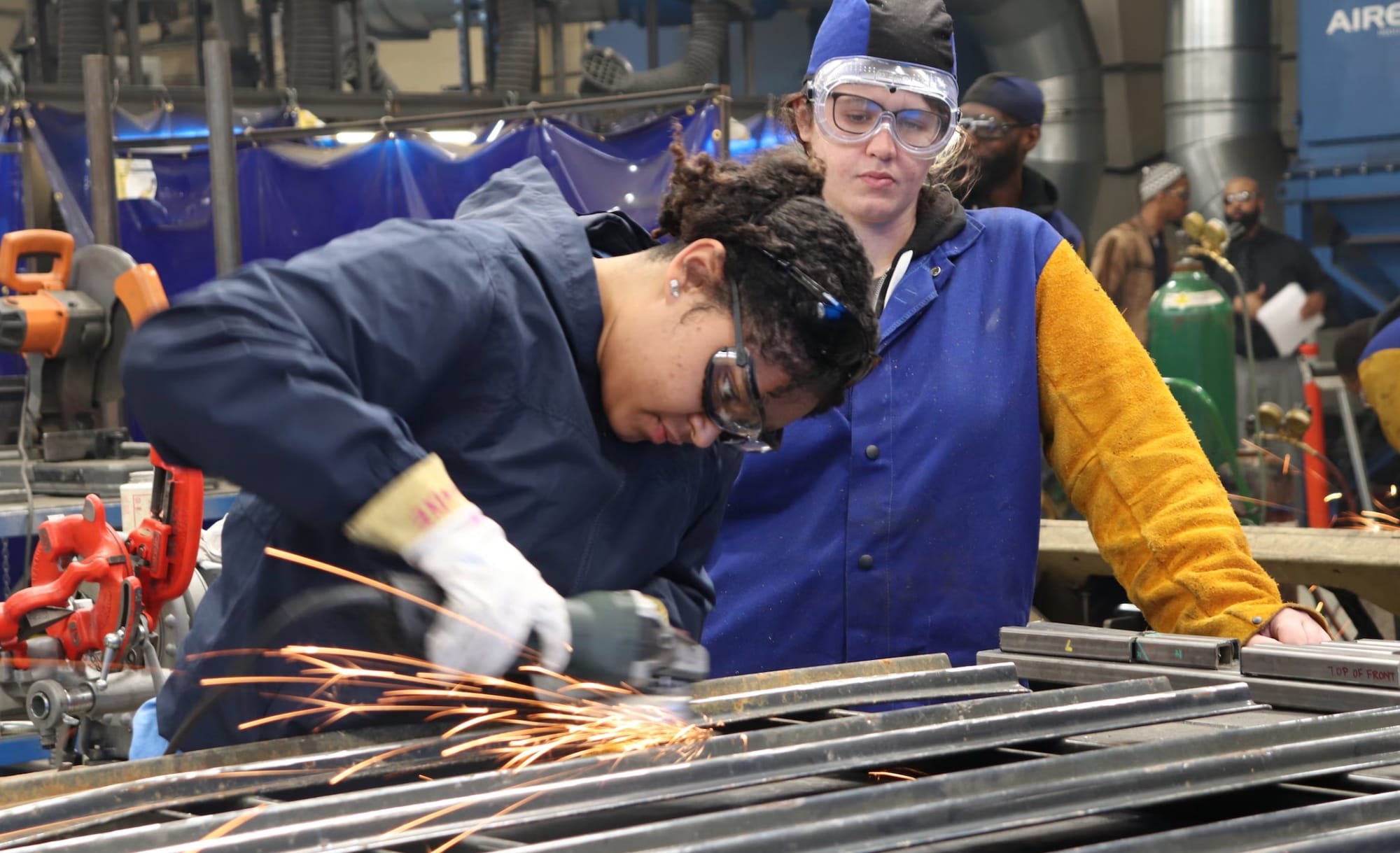 Woman cutting metal has dark hair & wears goggles & gloves. Supervisor wears goggles & a blue-and-gold jacket
