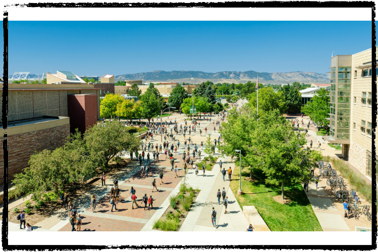 View to the mountains high up between two buildings. On the ground, many people walk between trees & grass