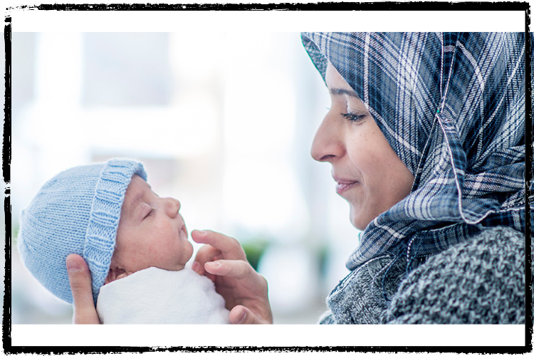 A woman wearing a blue, plaid hijab gently holds and gazes at her sleeping infant wearing a blue, knit beanie