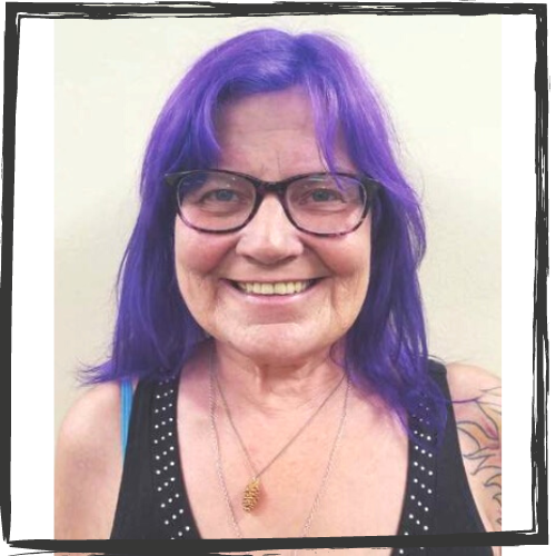 A white woman w/purple hair and glasses wears a black, sleeveless top and gold necklace