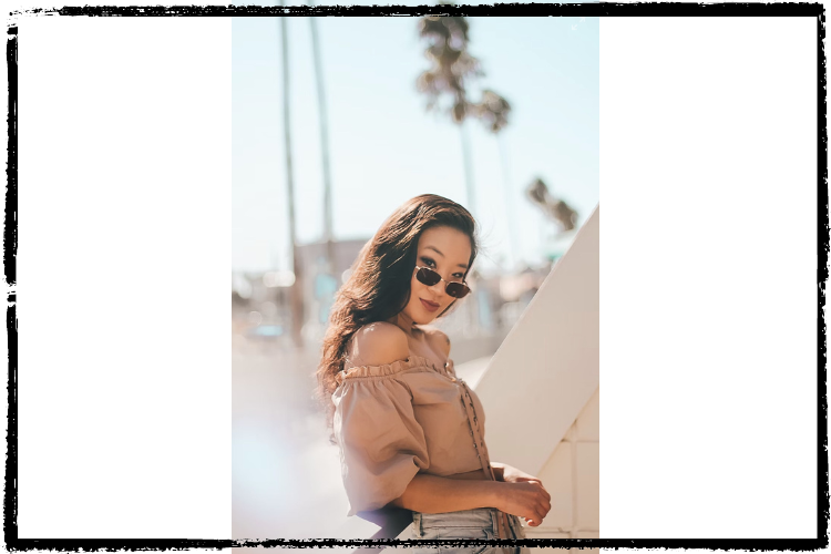 A young, Asian-looking woman wears oval sunglasses down her nose and an off-the-shoulder top; behind her are palm trees
