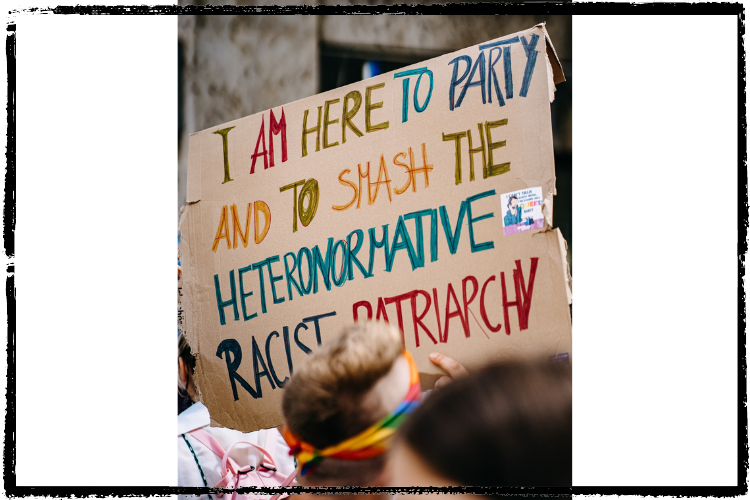 Cardboard sign w/colorful letters: "I am here to party and to smash the heteronormative racist patriarchy"