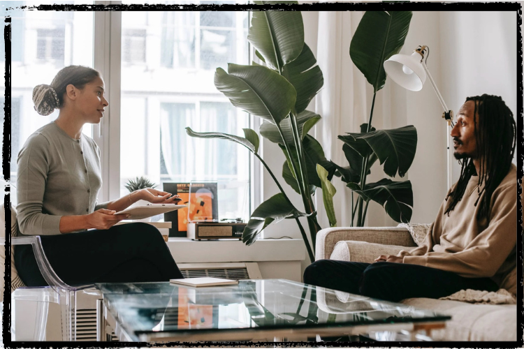A Woman of Color sits in a chair & talks to a Black man w/long braids sitting on a sofa 