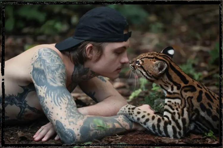 Photo of a young, white man laying on the ground balanced on his elbows and an ocelot lying next to him nose-to-nose
