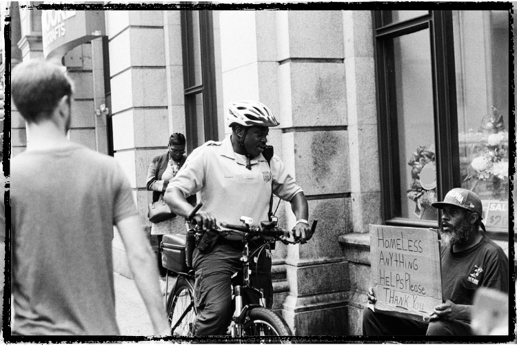 B&w photo of a bicycle cop talking to a homeless man holding a sign, "Homeless, Anything Helps Please, Thank You" 
