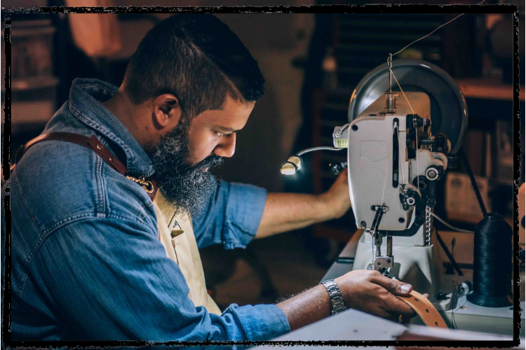 Photo of a brown man with short, black beard & fuzzy, black beard working with a leather belt at a sewing machine