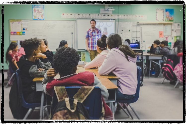 Photo of a classroom full of students facing away from the camera toward a teacher standing in front of a whiteboard talking