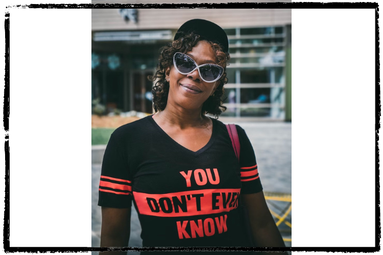 Photo of a Black woman smiling, wearing sunglasses and a black-and-red shirt that says, "You Don't Even Know"