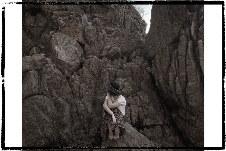 Photo of a woman wearing a dark hat & white shirt looking away and sitting on a rock surrounded by rocky walls