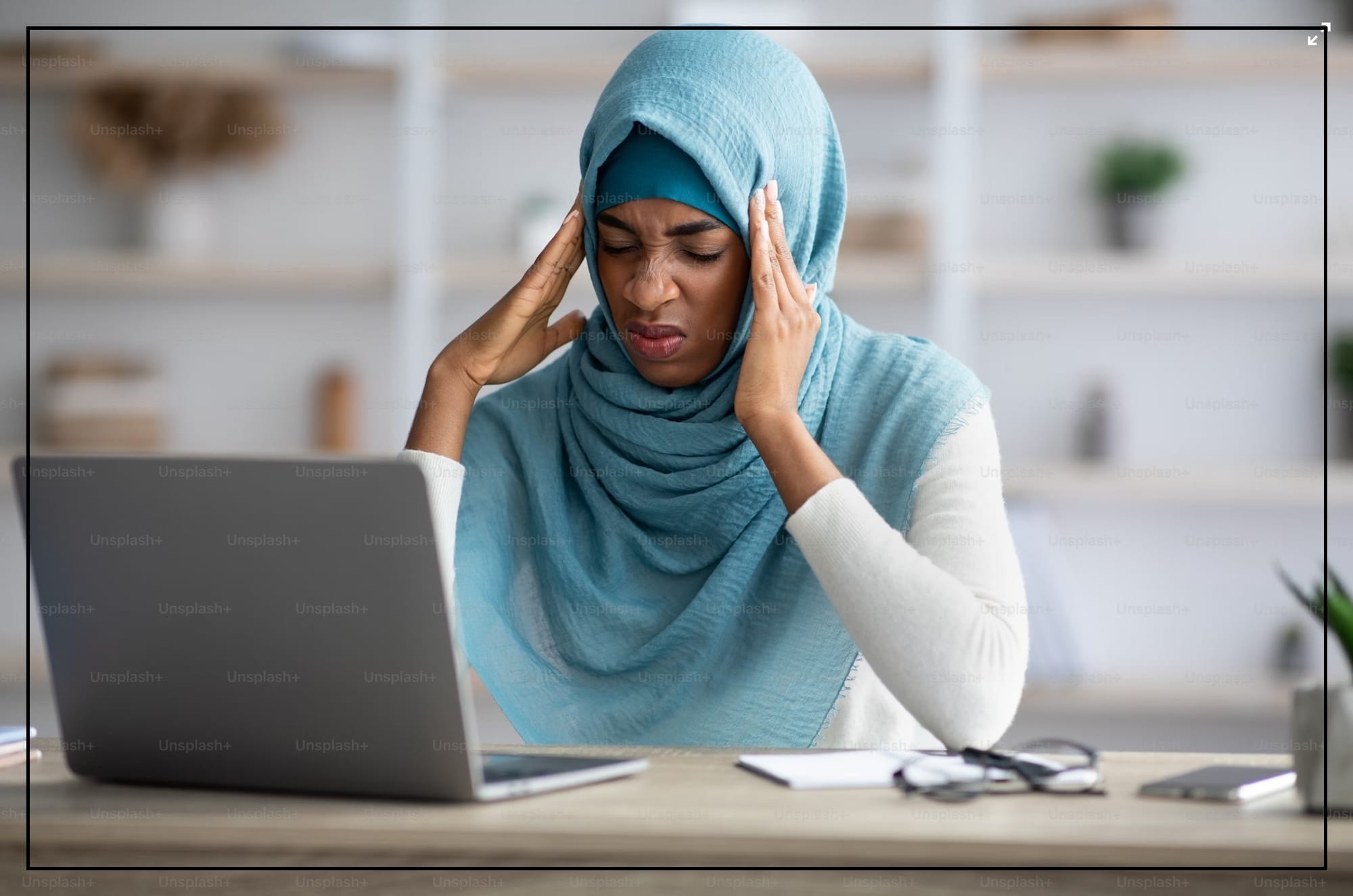 A woman in a hijab rubs her temples while sitting at a desk in front of a laptop