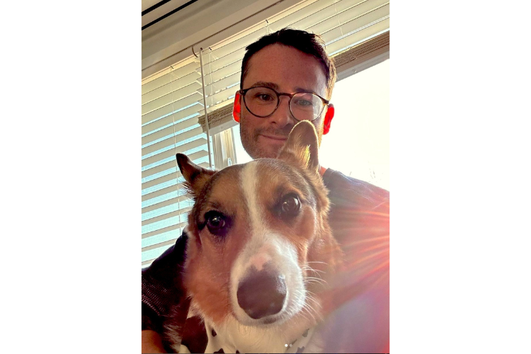 Photo of a white man with short, brown hair and wearing glasses next to his shepherd-mix dog