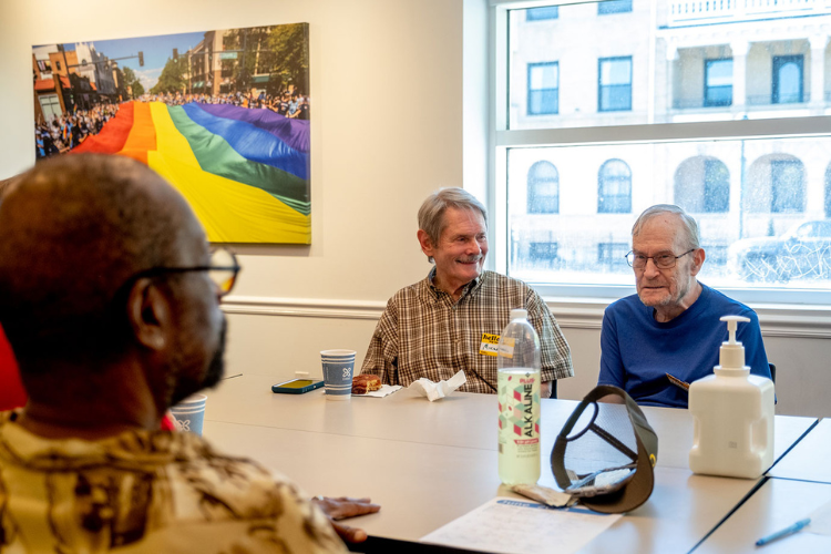 Photo of 2 elder white men sitting at a table facing the camera and a Black man on the other side facing away