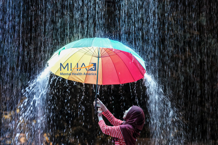 Photo of a woman in a hijab holding up a rainbow umbrella w/Mental Health America logo inside in the pouring rain. 