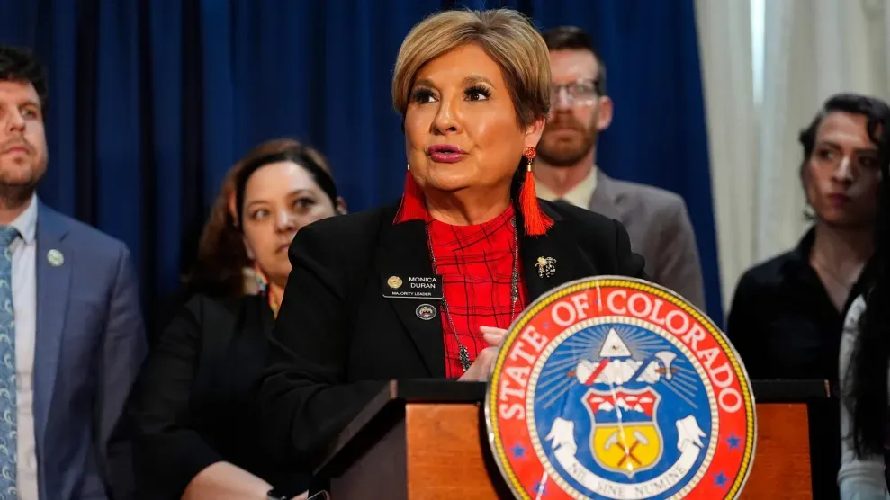 Monica Duran stand behind a lectern with the State of Colorado seal and surrounded by other people.