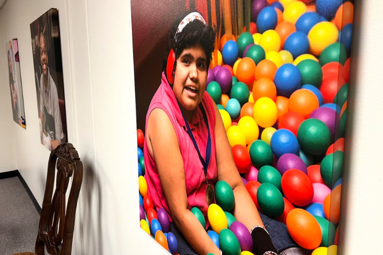 A young, Brown person with short, black hair wears headphones and sits in a colorful ball pit