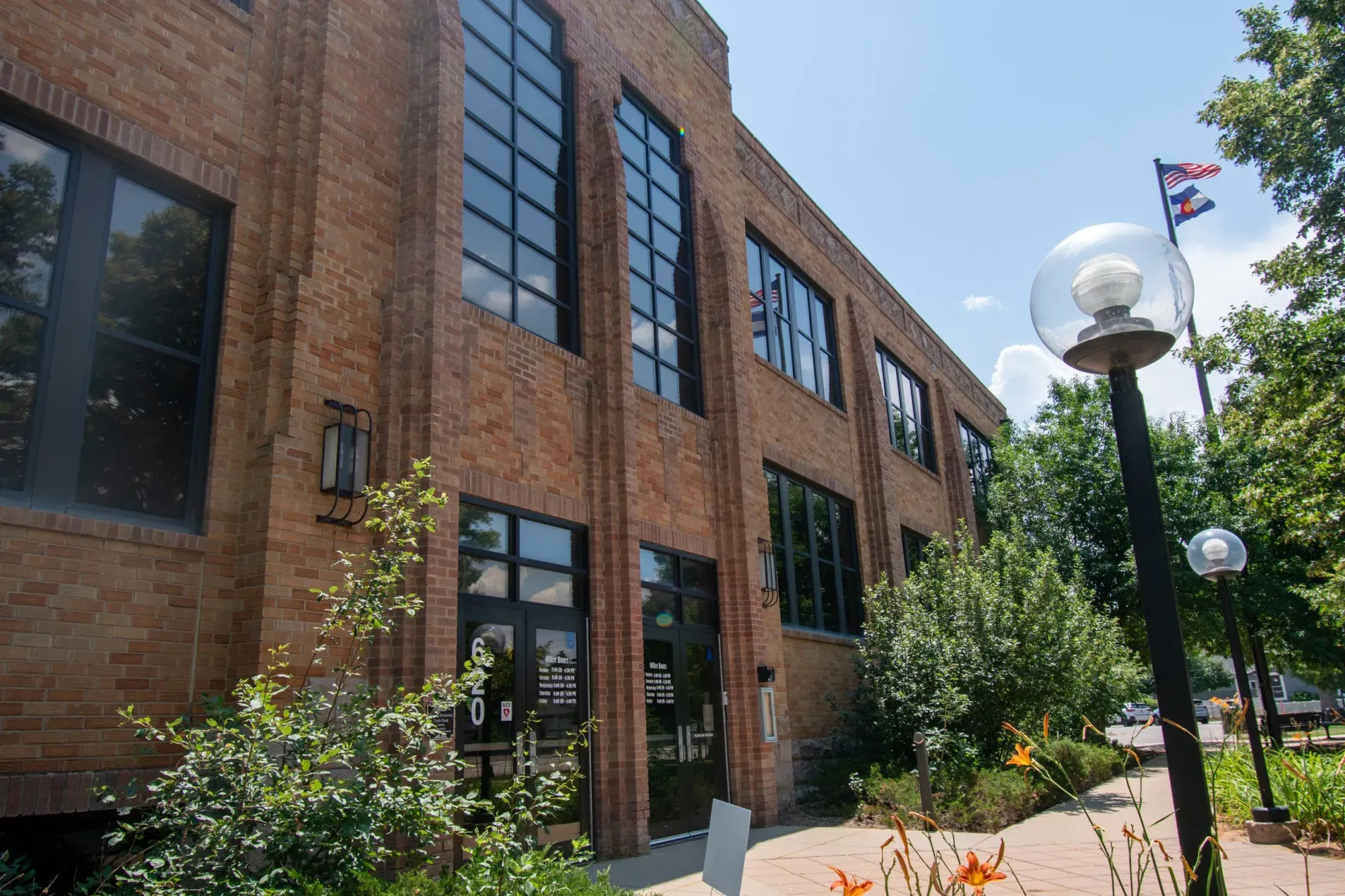 Photo of an orange brick building with dark framed windows. Shrubbery, trees and the American flag on a pole stand nearby