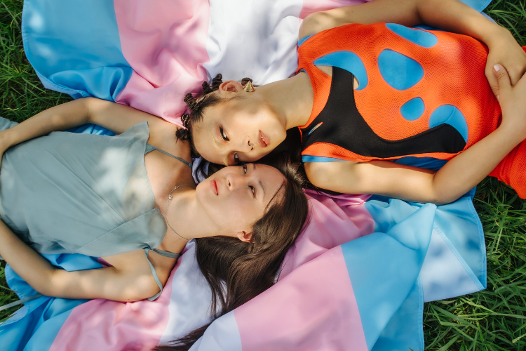 Two Trans people lie on a Trans flag in the grass, heads touching and bodies pointing in opposite directions