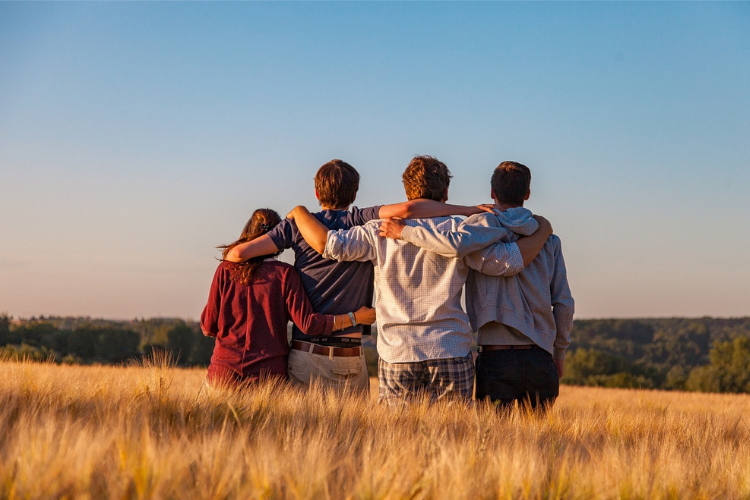 In a tall grass field, 4 people in a line hold each other looking away from the camera