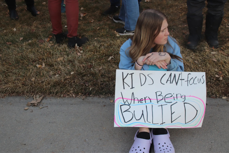 A white teen with long, brown hair sits on the grass holding a sign: "Kids can't focus when being bullied."