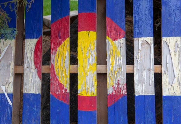 Fence with Colorado flag painted on it peeling away, fading and chipped in places