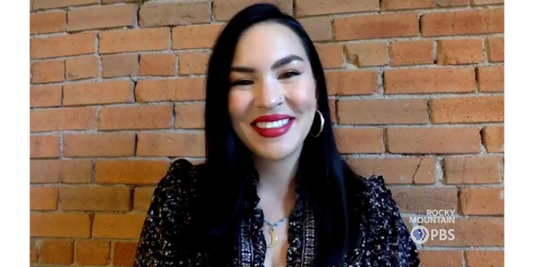 A Latina w/long hair and gold hoop earrings smiles at the camera. She wears red lipstick and a black, floral blouse