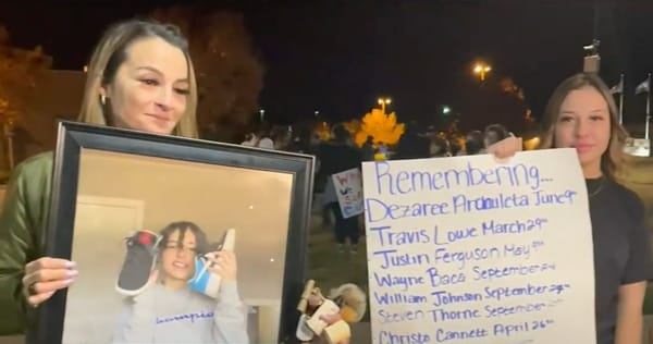 Photo of two women w/long hair. 1 holds a portrait of her daughter. 1 holds a sign that begins, "Remembering..."