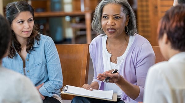 A middle-aged Black woman w/a clipboard speaks to a women's group. A Latina and others listen