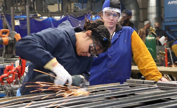 Woman cutting metal has dark hair & wears goggles & gloves. Supervisor wears goggles & a blue-and-gold jacket