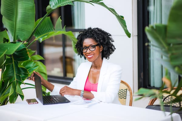 A Black woman with curly, black hair wears glasses and a white suit with pink shirt. She is smiling