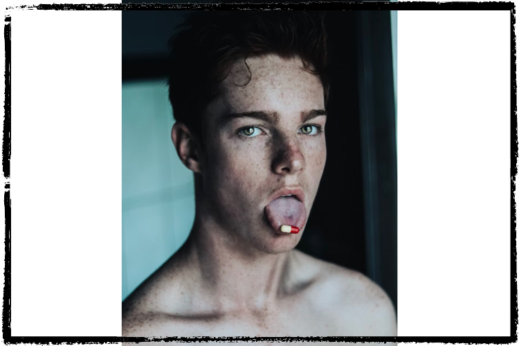 A young, white man w/short, curly hair holds a red-and-white pill on the end of his tongue