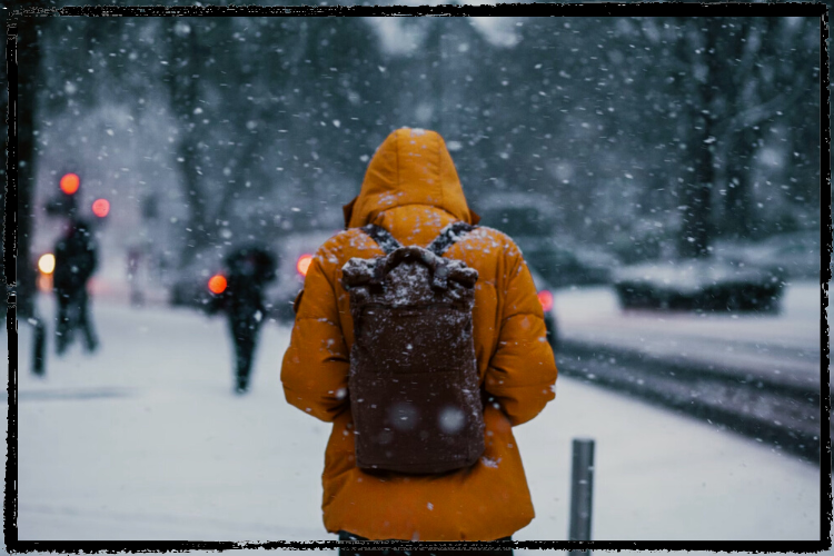 Photo of person bundled in an orange winter coat and carrying a backpack walking away from camera as it snows