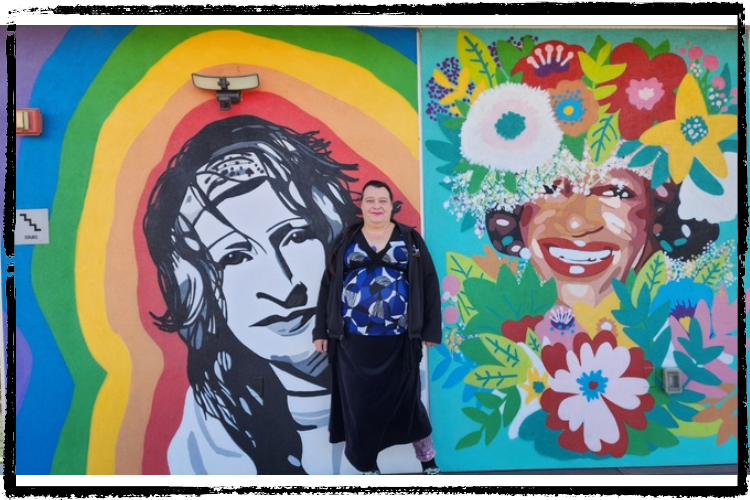 A woman w/short, black hair wearing a colorful blouse & black skirt stands in front of 2 side-by-side colorful murals of wome