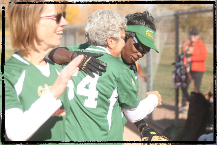Photo of 3 women in green-and-white softball jerseys hugging each other and smiling