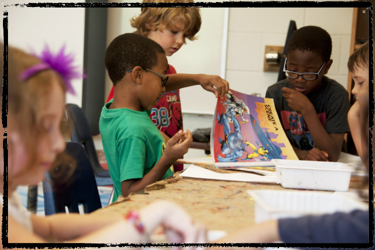 Photo of 5 diverse, young students looking at documents on a table or through a colorful book 