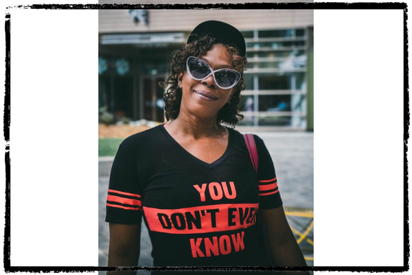 Photo of a Black woman smiling, wearing sunglasses and a black-and-red shirt that says, "You Don't Even Know"