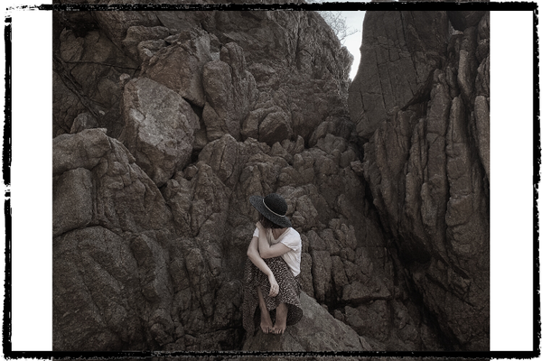 Photo of a woman wearing a dark hat & white shirt looking away and sitting on a rock surrounded by rocky walls