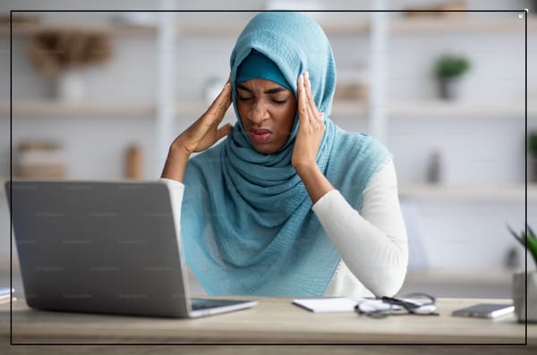 A woman in a hijab rubs her temples while sitting at a desk in front of a laptop