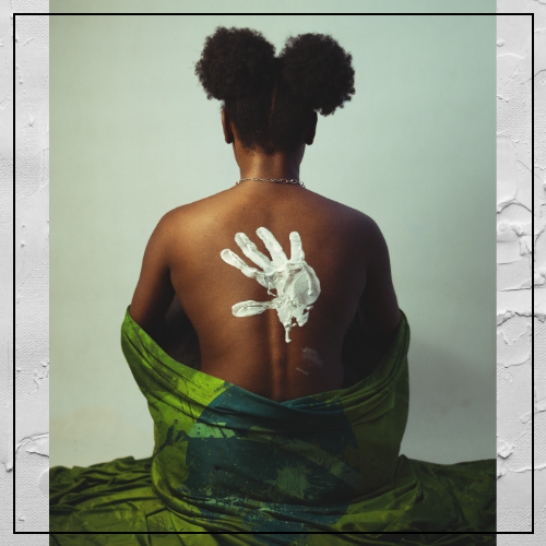 A black woman wearing a green dress sits with her back to the camera; on her bare back is a white handprint